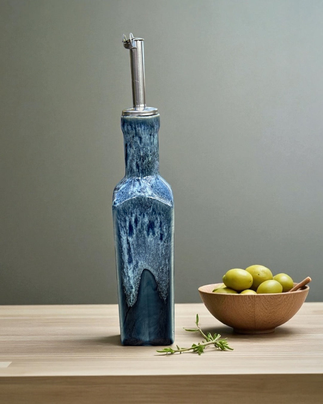 Handmade Ceramic Olive Oil Bottle in deep blue glaze with stainless steel pourer, accompanied by a bowl of green olives.