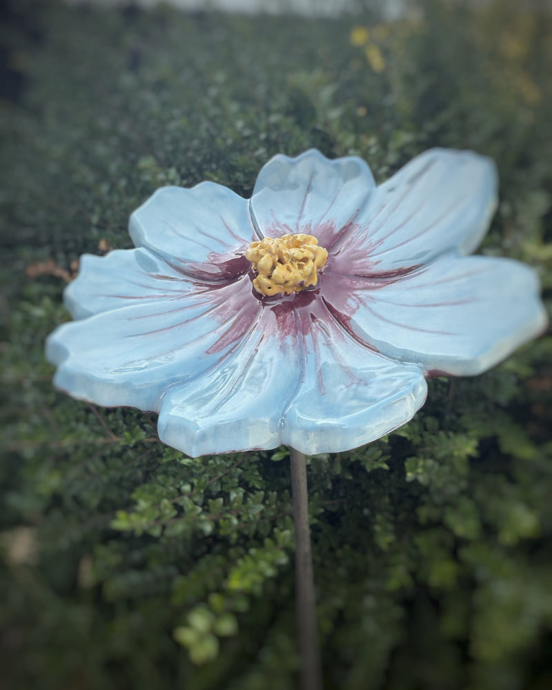 Outdoor handmade ceramic flower in blue and purple, featuring a yellow center, surrounded by greenery.
