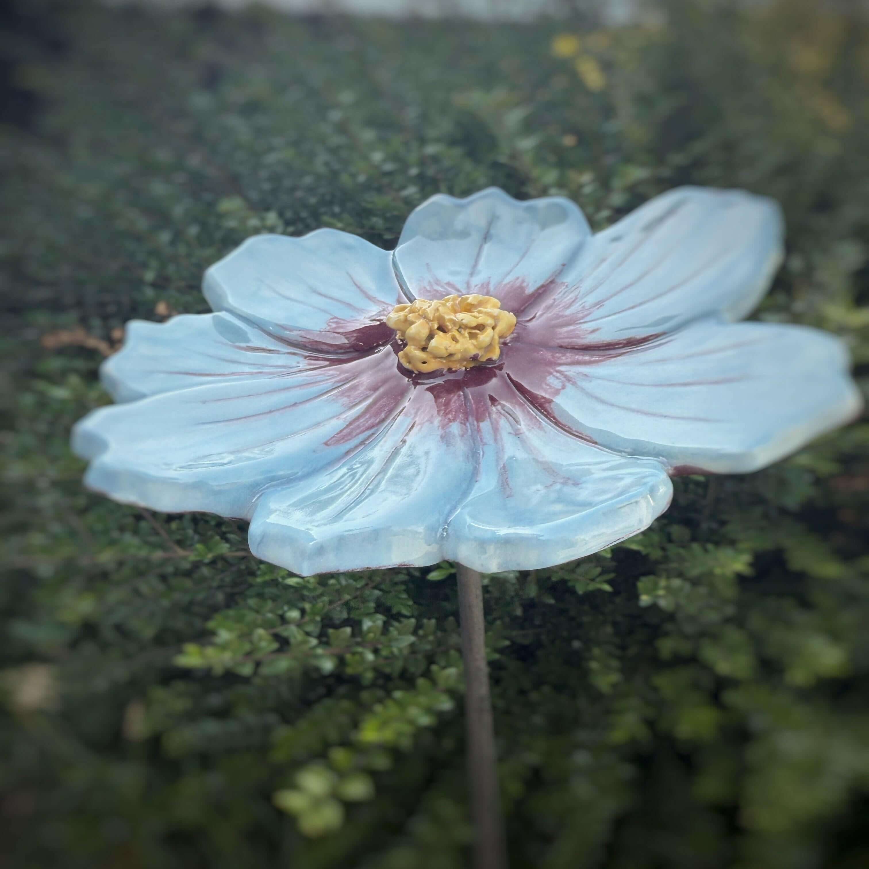 Handmade ceramic garden flower in sky-blue with purple accents and yellow center on a garden stake.