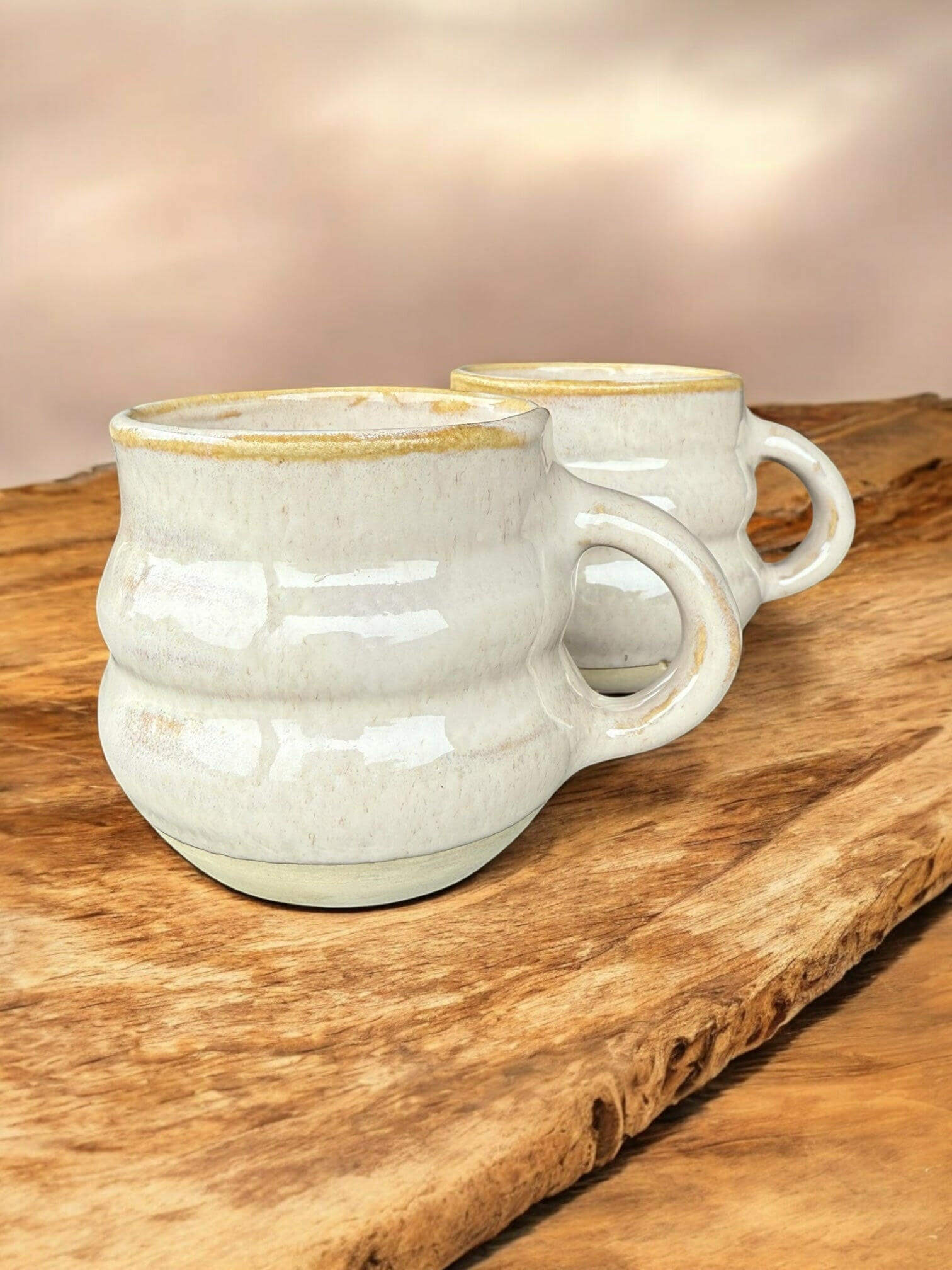 Two handmade ribbed mugs in speckled stoneware with glossy white glaze and earthy rims on a rustic wooden surface.
