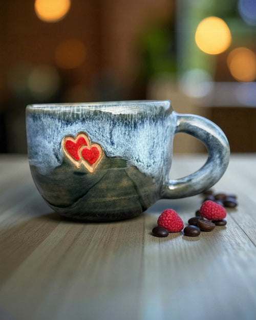 Blue glazed stoneware mug with hearts on a wooden table with coffee beans and raspberries.