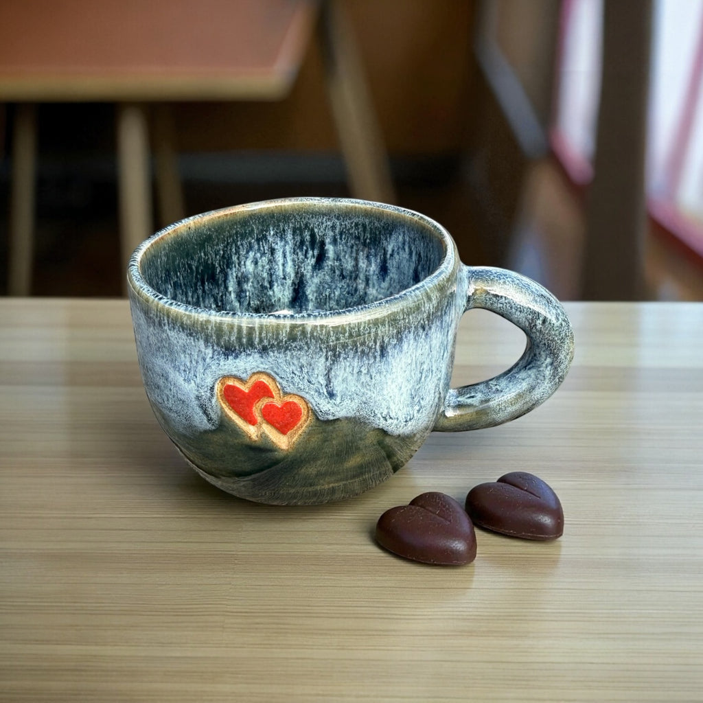 Blue glazed stoneware mug with hearts beside two chocolate hearts on a wooden table.