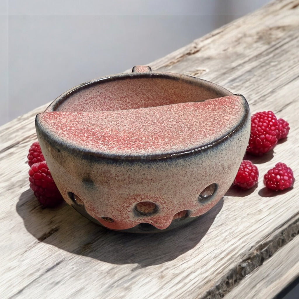 Ceramic berry rinsing mug with soft strawberry glaze, surrounded by fresh raspberries on a wooden surface.
